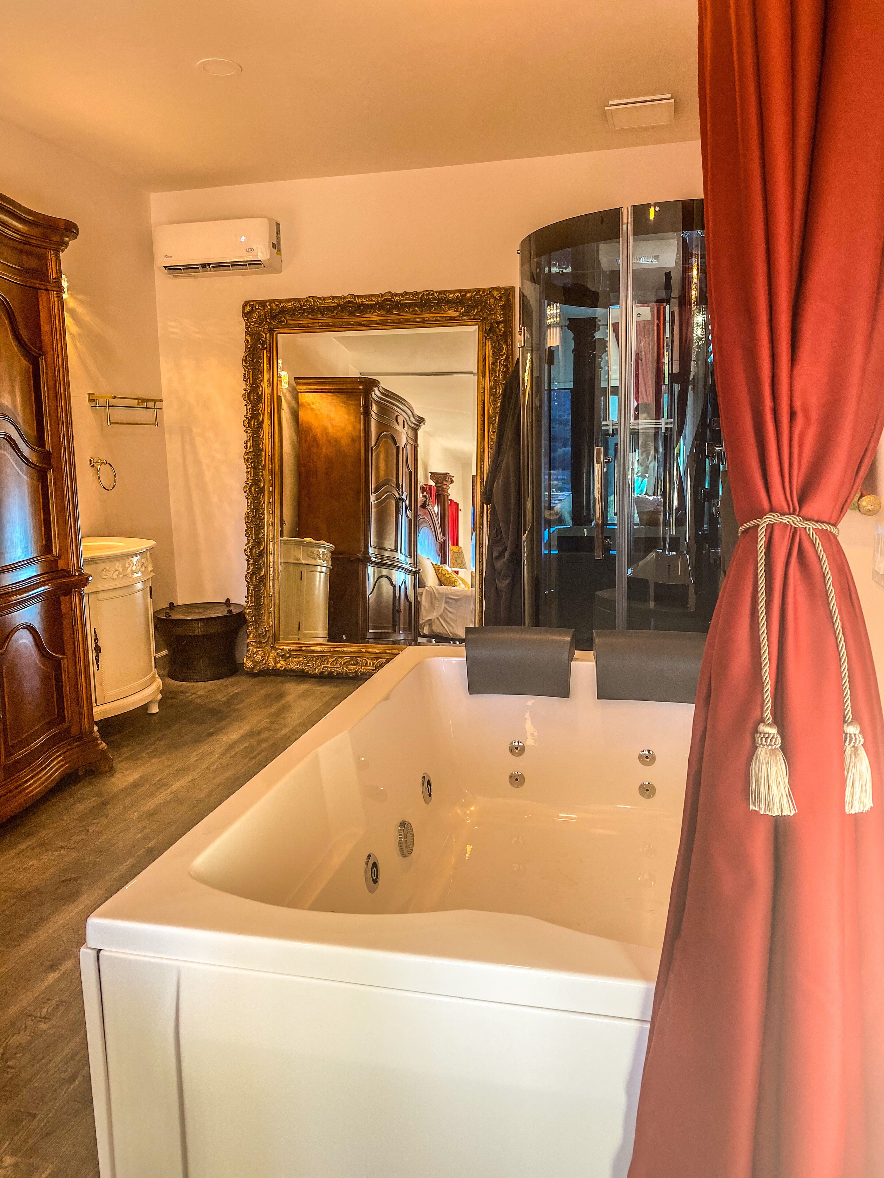 Una habitación con una bañera blanca grande, un armario de madera, un espejo con un marco decorado y una cortina roja.
