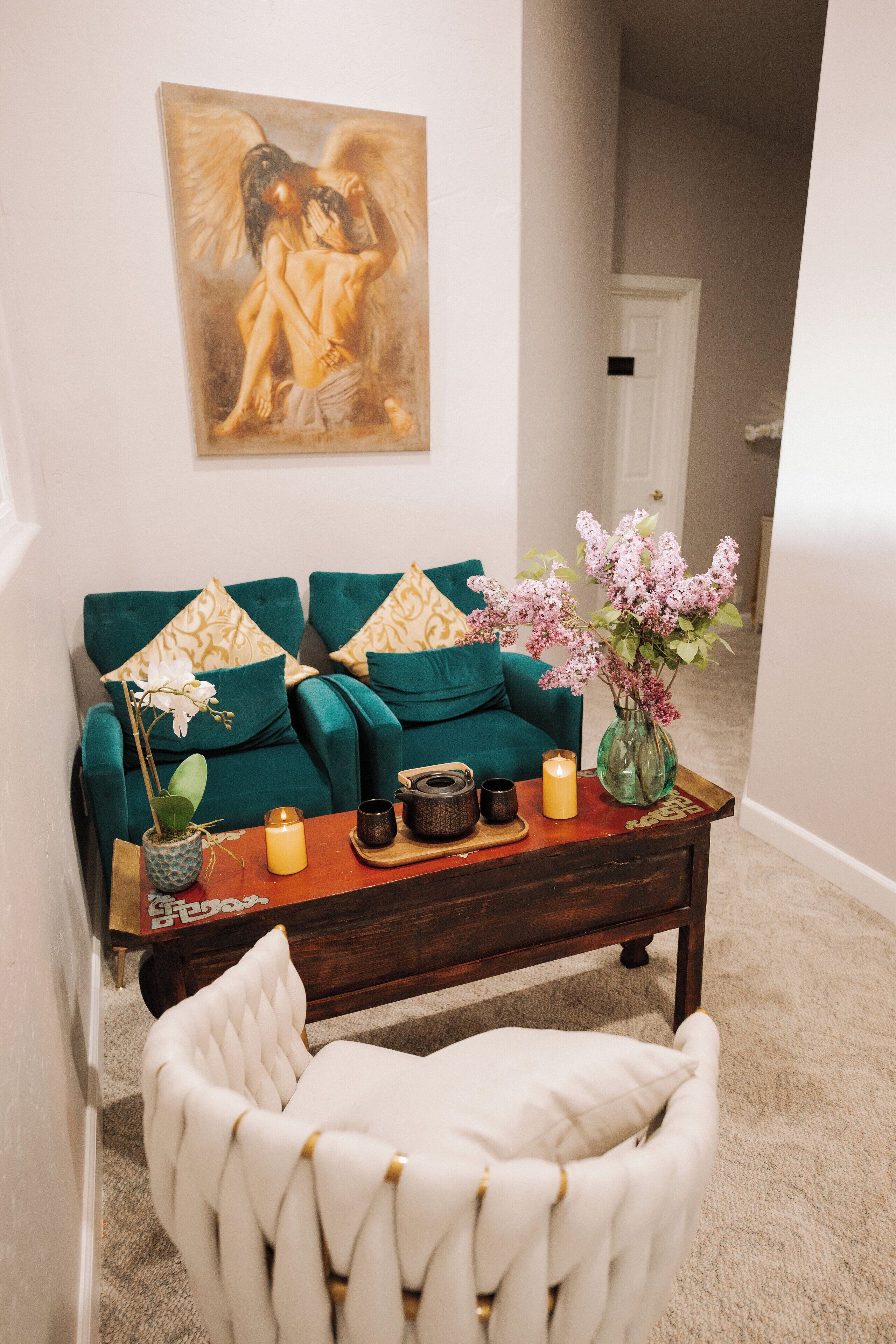 A living room with a teal sofa, a white armchair, a coffee table with decorative items, and a painting on the wall.