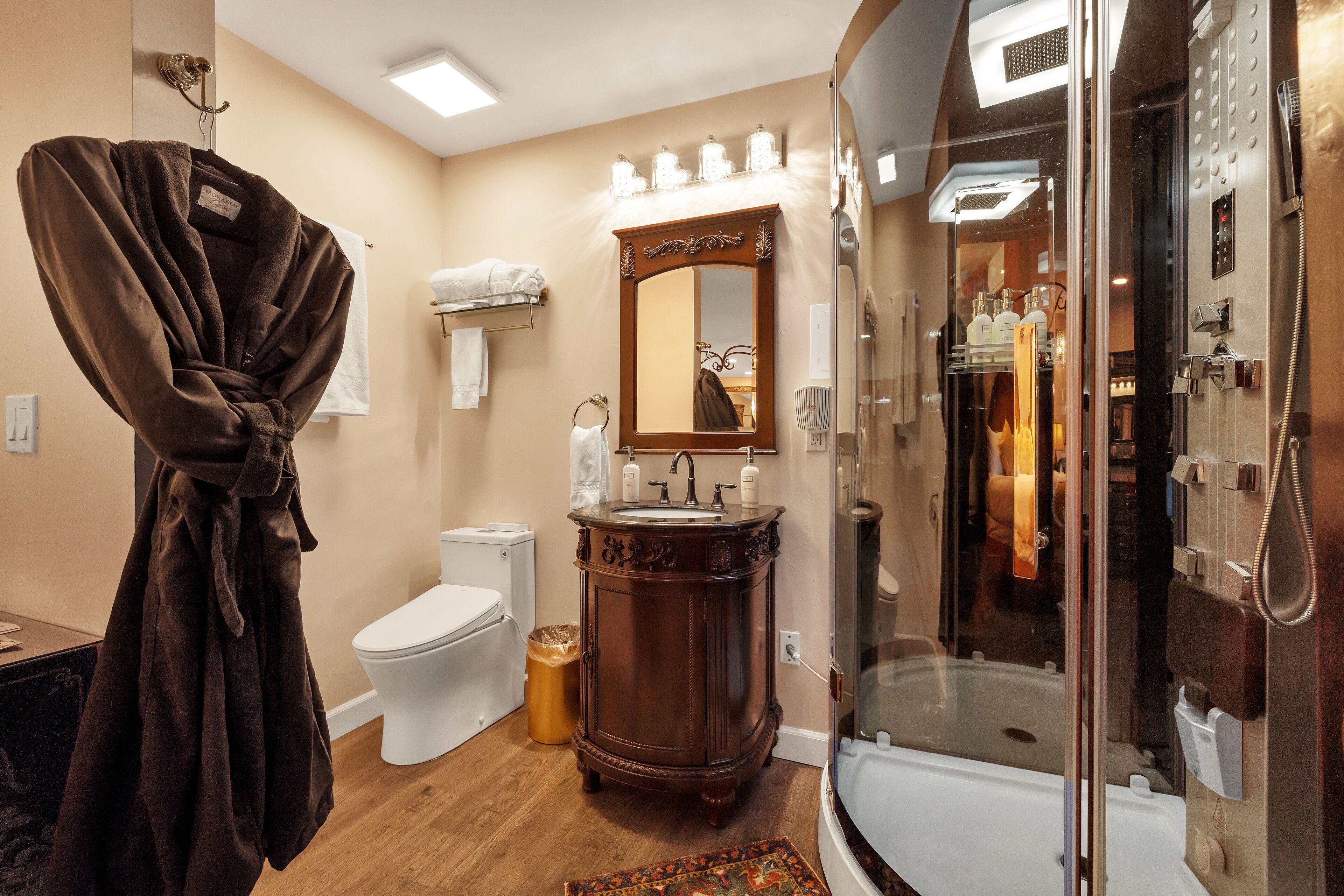 Une salle de bain avec une douche à parois de verre, un meuble-lavabo en bois, des toilettes blanches et une robe suspendue à un crochet.