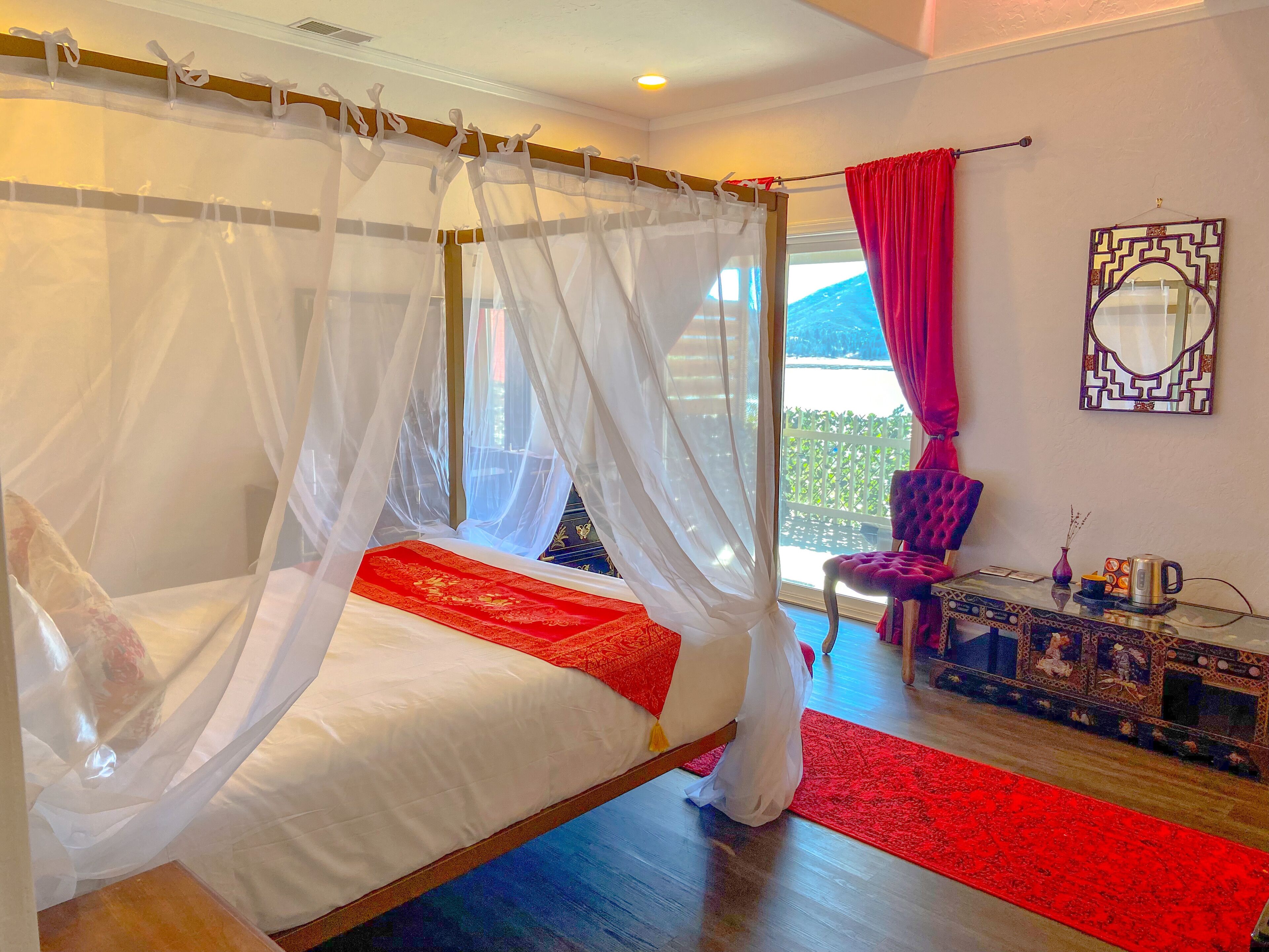 Una cama con dosel, ropa de cama blanca y un corredor decorativo rojo, una alfombra estampada en rojo y negro, un sillón púrpura y un espejo decorativo en la pared.