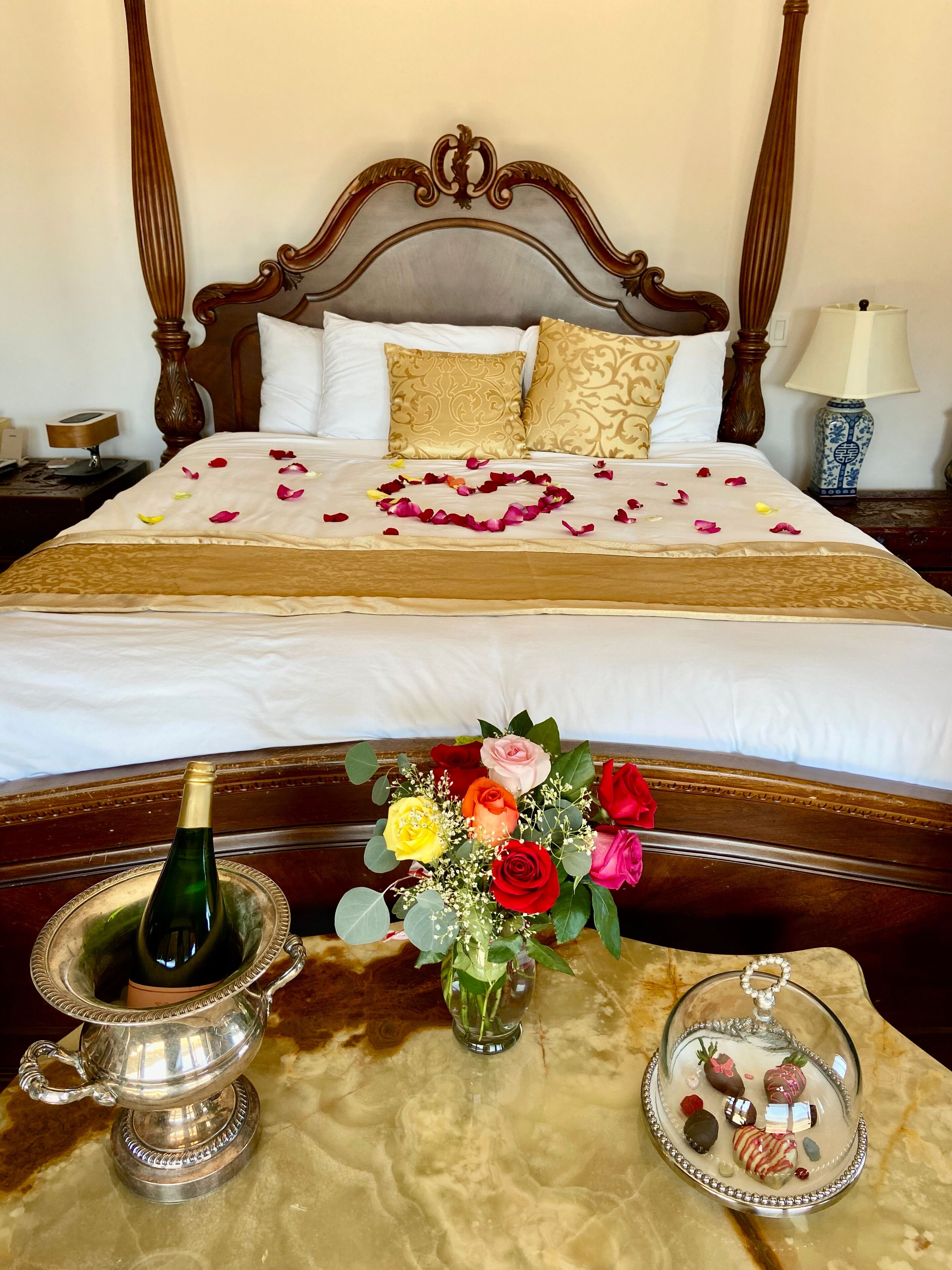 Een netjes opgemaakt bed met een decoratief hoofdbord, een nachtkastje met een lamp, en een dienblad met chocolade en een fles champagne.