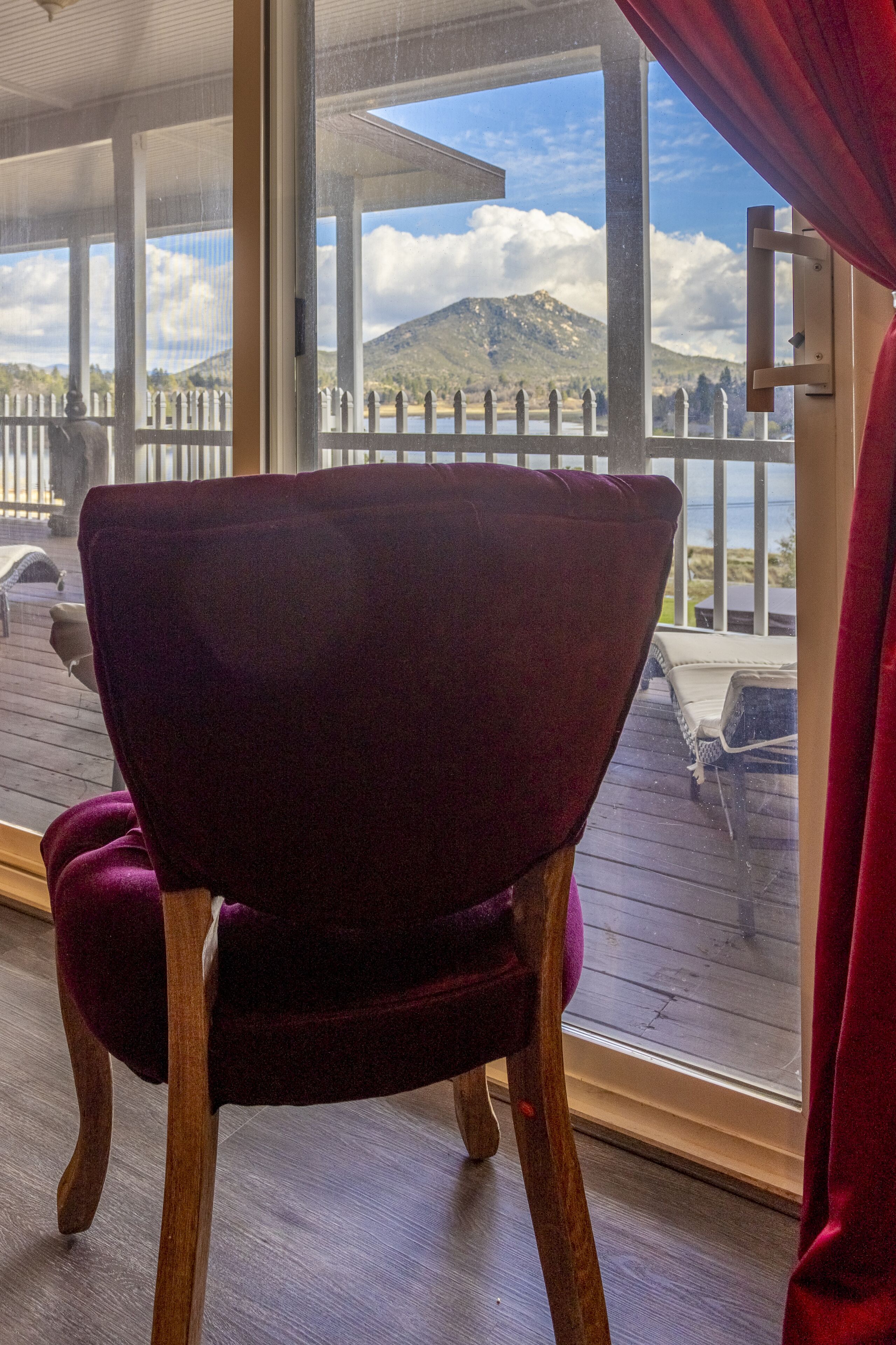 Una silla de madera con cojín, ubicada cerca de una ventana con vista a una terraza con cerca blanca y una montaña al fondo.