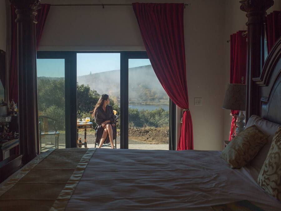 Ein Schlafzimmer mit einem großen Bett, Blick ins Freie durch eine Schiebetür und einem Tisch mit einer Person draußen.