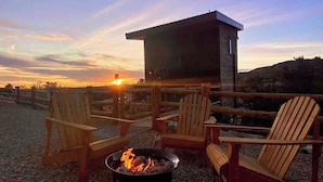 A fire pit with chairs and a small building, likely a cabin, with a sunset in the background.