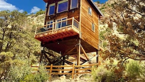 A wooden cabin with a balcony, situated on stilts amidst a rocky landscape with trees.