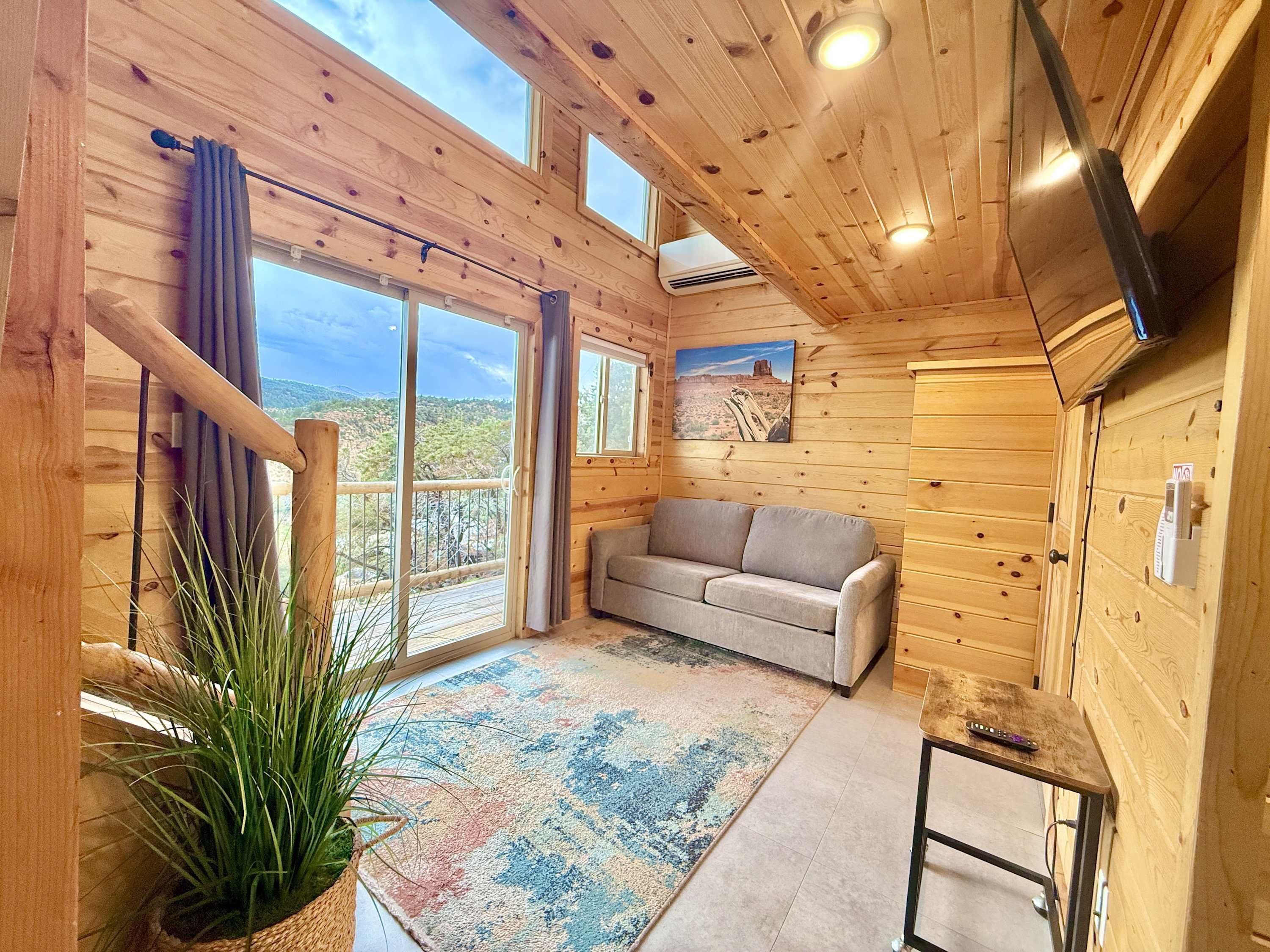 A cozy wooden cabin interior with a sofa, a large window, and a potted plant.