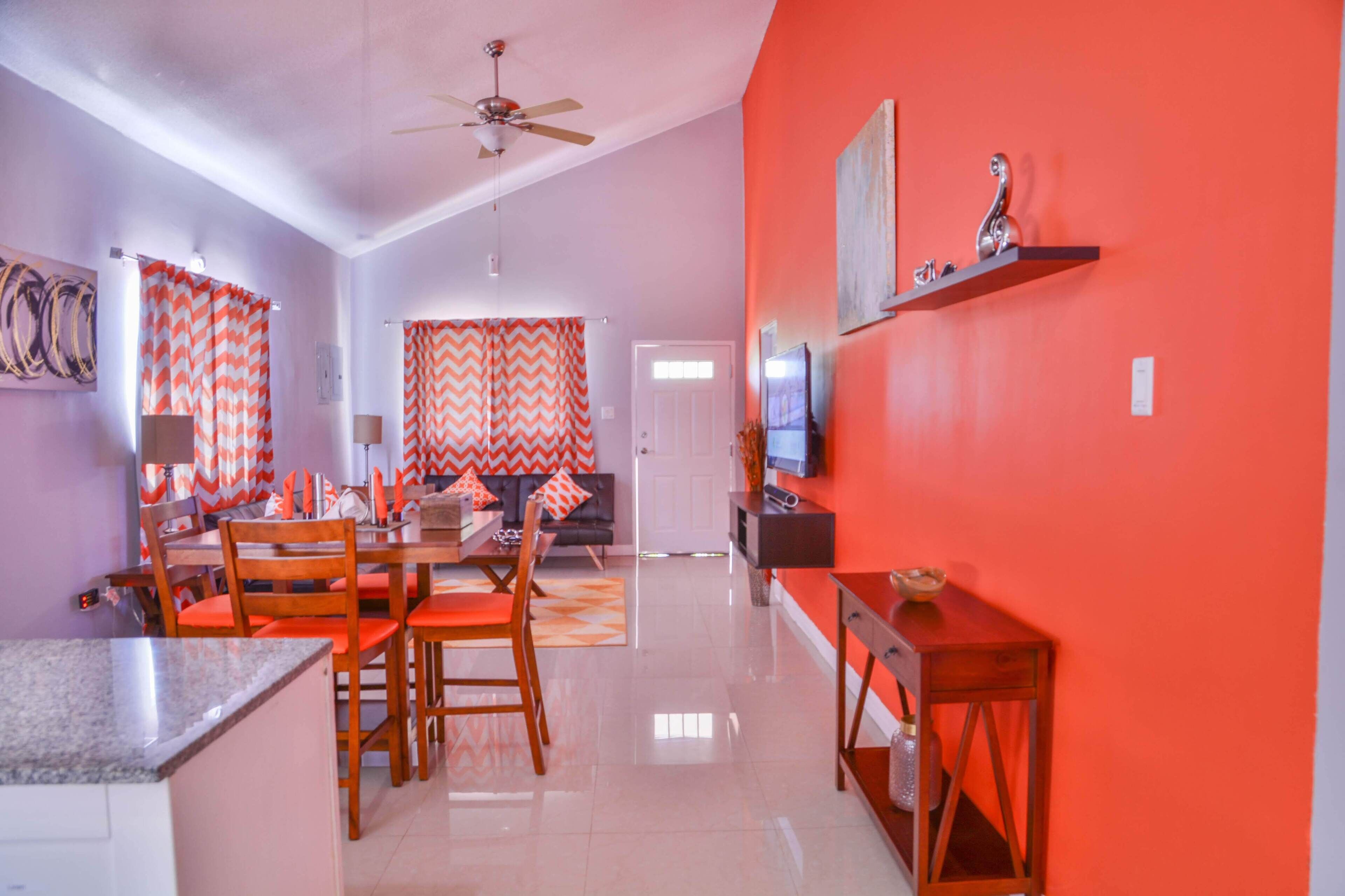 A modern living room with a sofa, dining table, and chairs. There is a television on a stand and a ceiling fan.