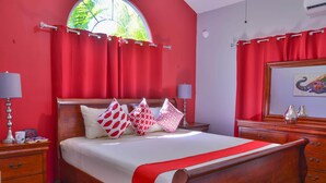Een slaapkamer met een groot bed, rode gordijnen, een houten hoofdbord, twee nachtkastjes met lampen en een ingelijst schilderij aan de muur.