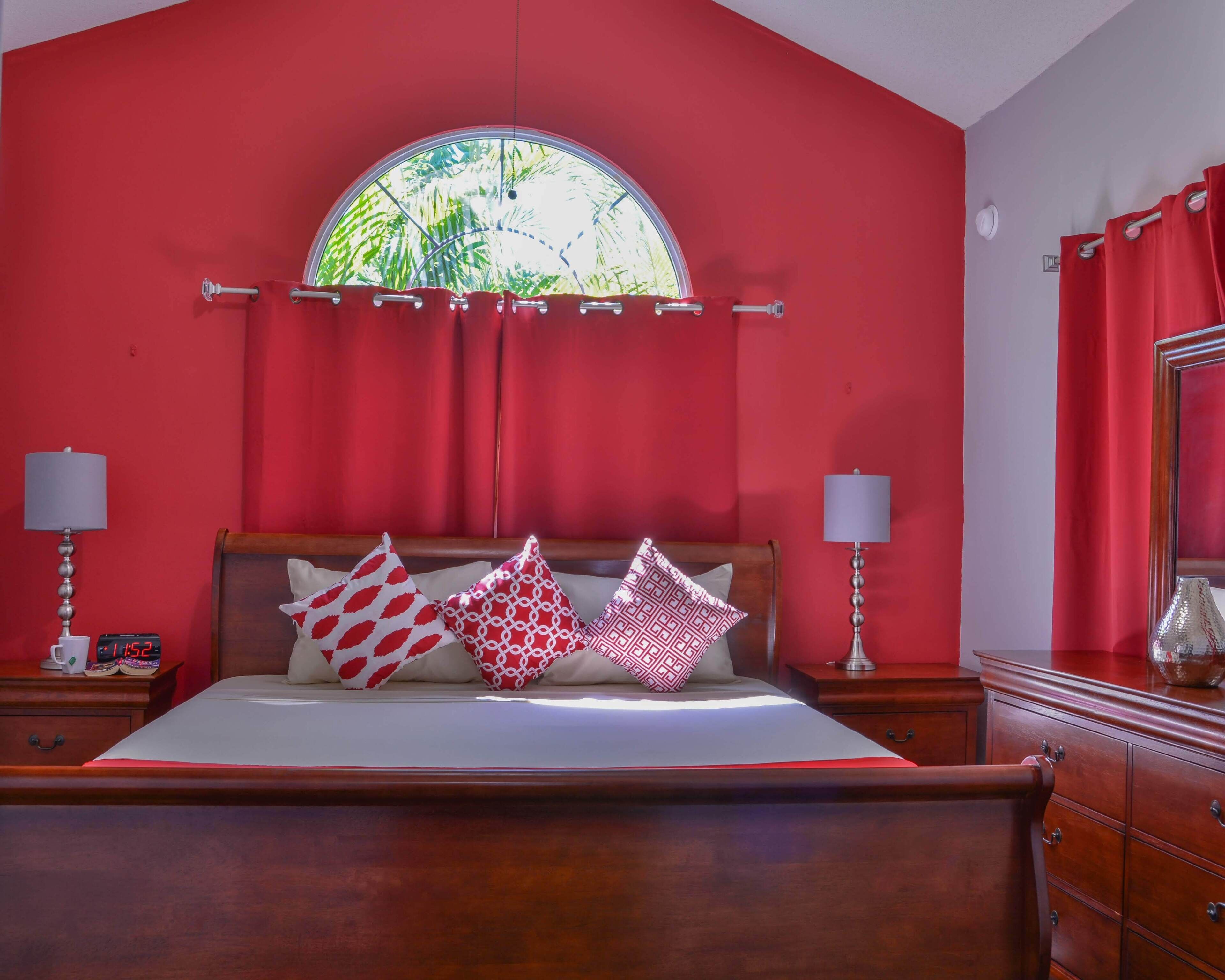 A bedroom with a large bed, red walls, a wooden headboard, and a window with a view of greenery.