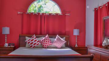 A bedroom with a large bed, red walls, a wooden headboard, and a window with a view of greenery.