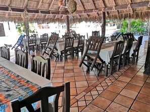 An outdoor restaurant with wooden tables and chairs, a tiled floor, and a thatched roof.