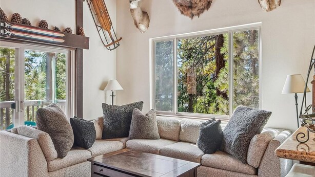Un salon chaleureux avec un canapé d’angle, une table basse et une cheminée. Des têtes d’animaux empaillées sont accrochées au mur, et une fenêtre offre une vue sur les arbres.