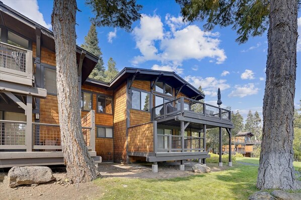 A modern wooden cabin with a balcony, surrounded by trees and a clear sky.