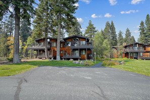 A two-story log cabin with a balcony, surrounded by tall trees and a paved driveway.