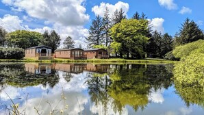 Una cabaña junto al lago, con su reflejo en el agua, rodeada de árboles y un cielo despejado.