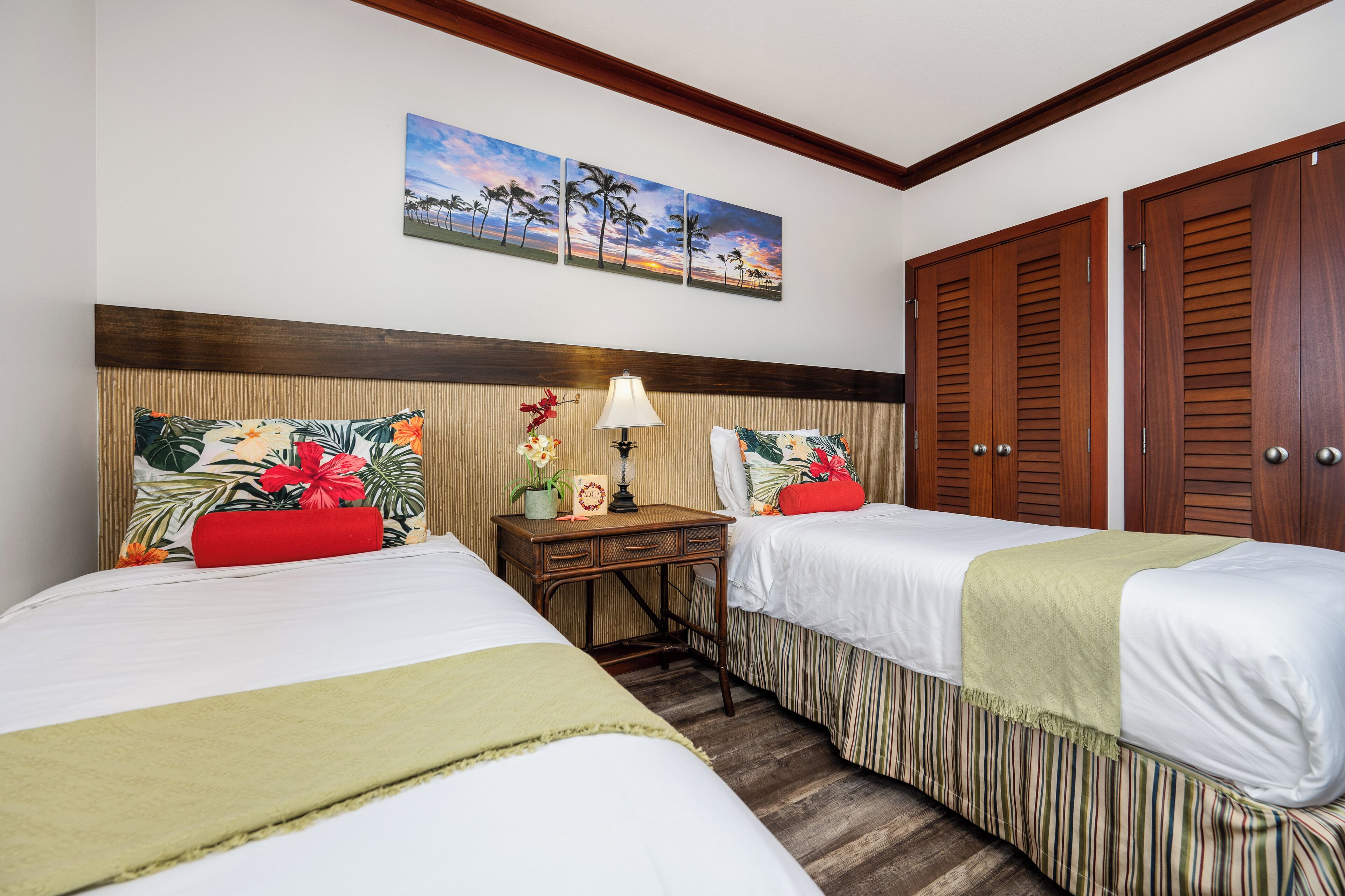 Habitación de hotel con dos camas, cabecera de madera, mesita de noche con lámpara y un cuadro de una escena tropical.