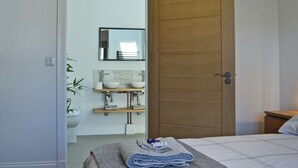 Una camera da letto con una porta in legno, un letto con copriletto bianco e grigio, un comodino e un bagno con lavandino e specchio.