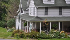Une maison à deux étages avec un porche, entourée d’un jardin où poussent différentes plantes et fleurs.