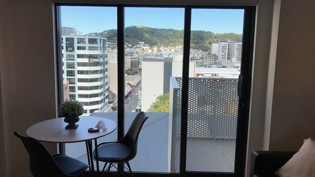Una habitación moderna con un amplio ventanal que ofrece vistas a la ciudad, una mesa redonda con dos sillas y un sofá negro con un cojín.
