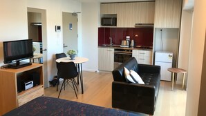 Un espacio habitable compacto con una cama, un sofá de cuero negro, una mesa de comedor con dos sillas, una pequeña cocina con microondas y refrigerador, y un televisor sobre un pedestal de madera.