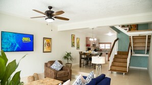 Un vaste espace de vie doté d’un ventilateur de plafond, d’un téléviseur diffusant la chaîne « Jamaica Travel Channel », d’un coin repas et d’un escalier menant à un niveau supérieur.