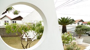 Une vue à travers une fenêtre circulaire sur un quartier résidentiel, avec des maisons, un palmier et un mur blanc.
