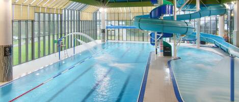 An indoor swimming pool with multiple lanes, a water slide, and a glass-enclosed area.