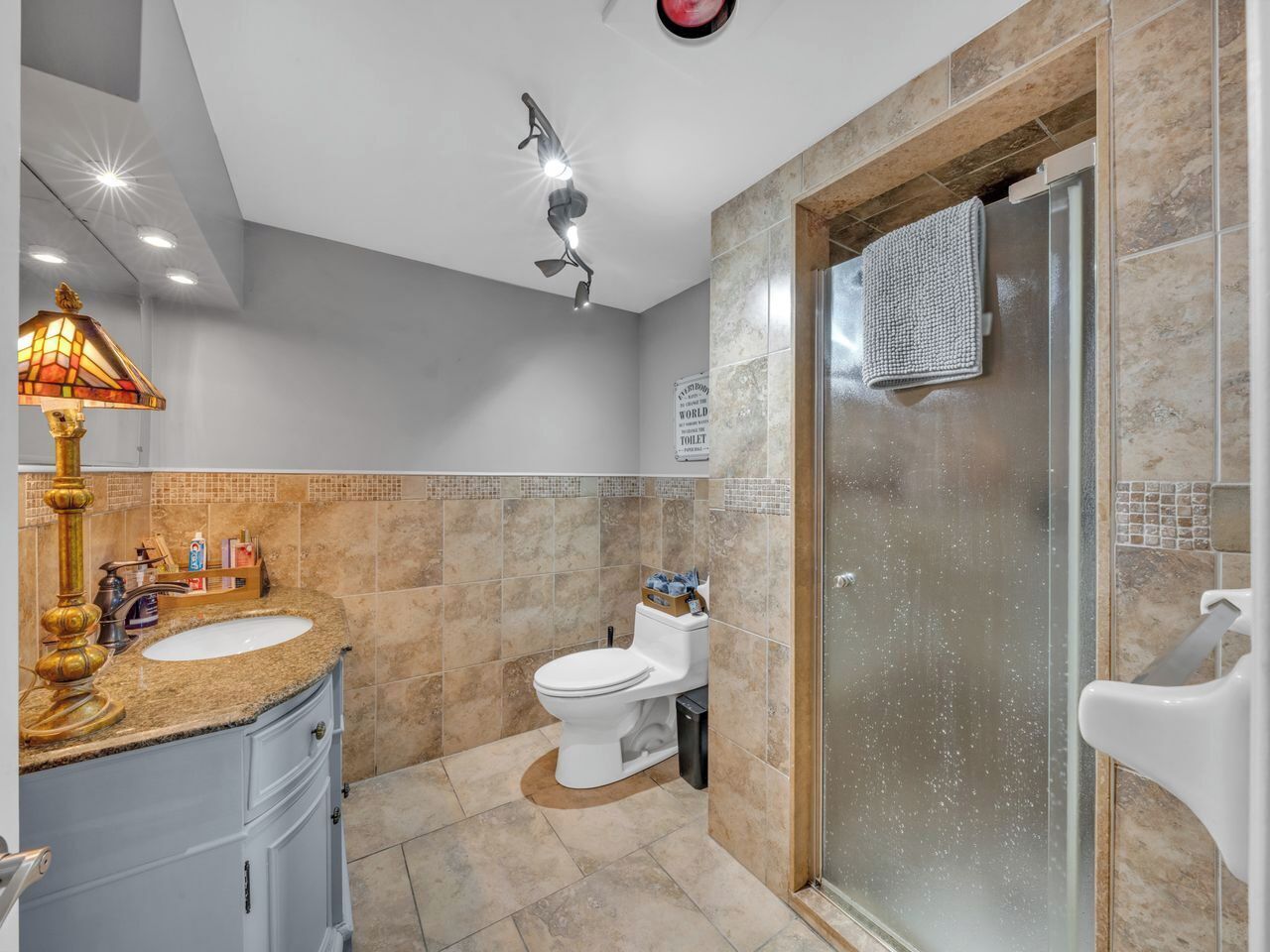 A modern bathroom with a granite countertop, a white toilet, a glass shower enclosure, and a decorative lamp.