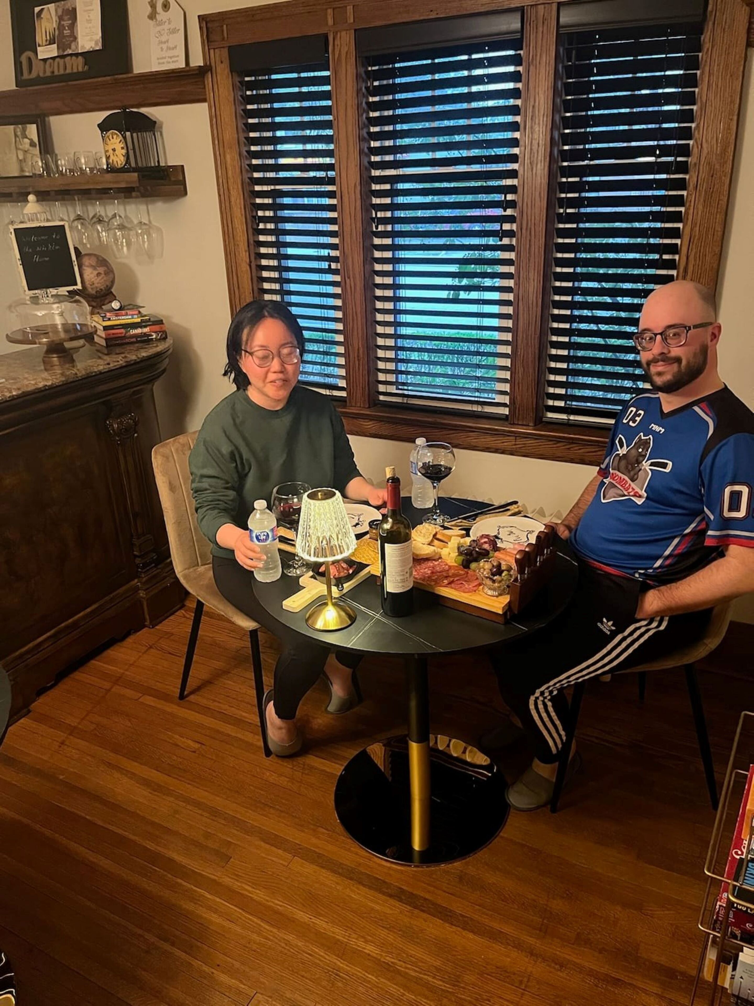 A dining table with a variety of food and drinks, a clock, and a window with blinds.