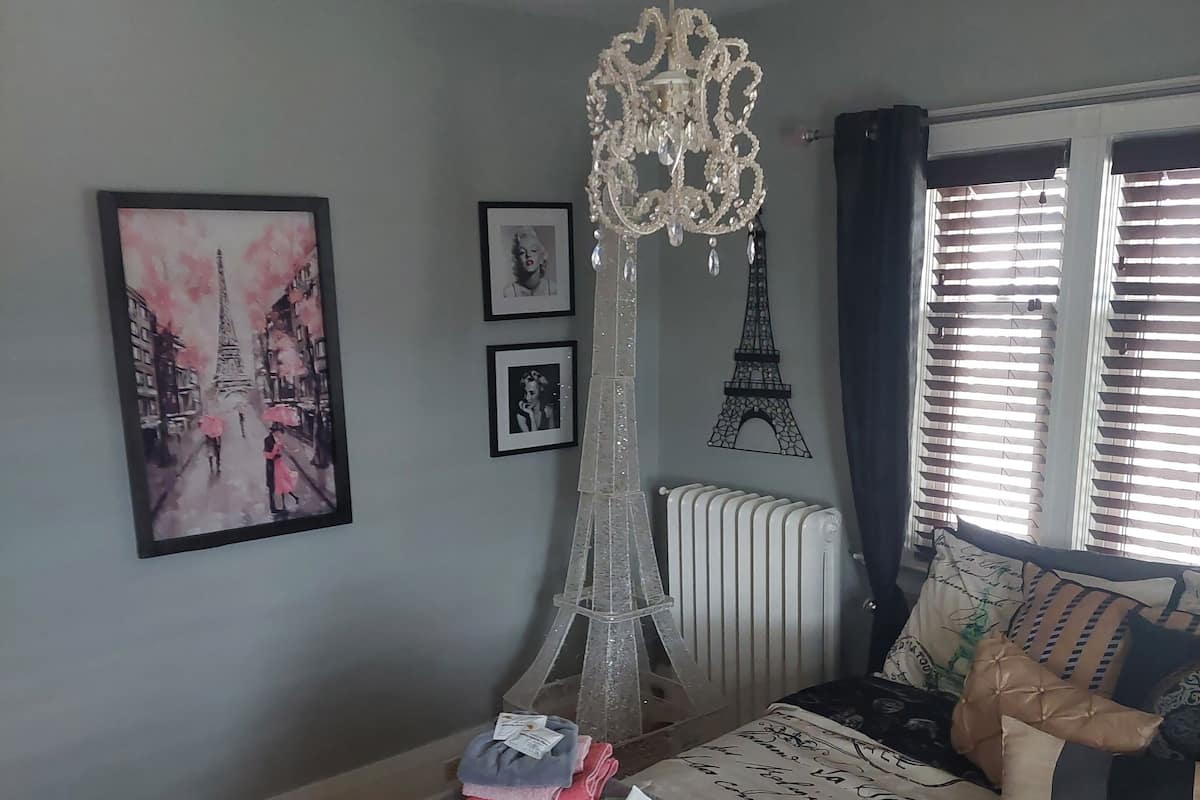 A bedroom with a bed featuring Eiffel Tower prints, a chandelier, and a stack of folded towels.