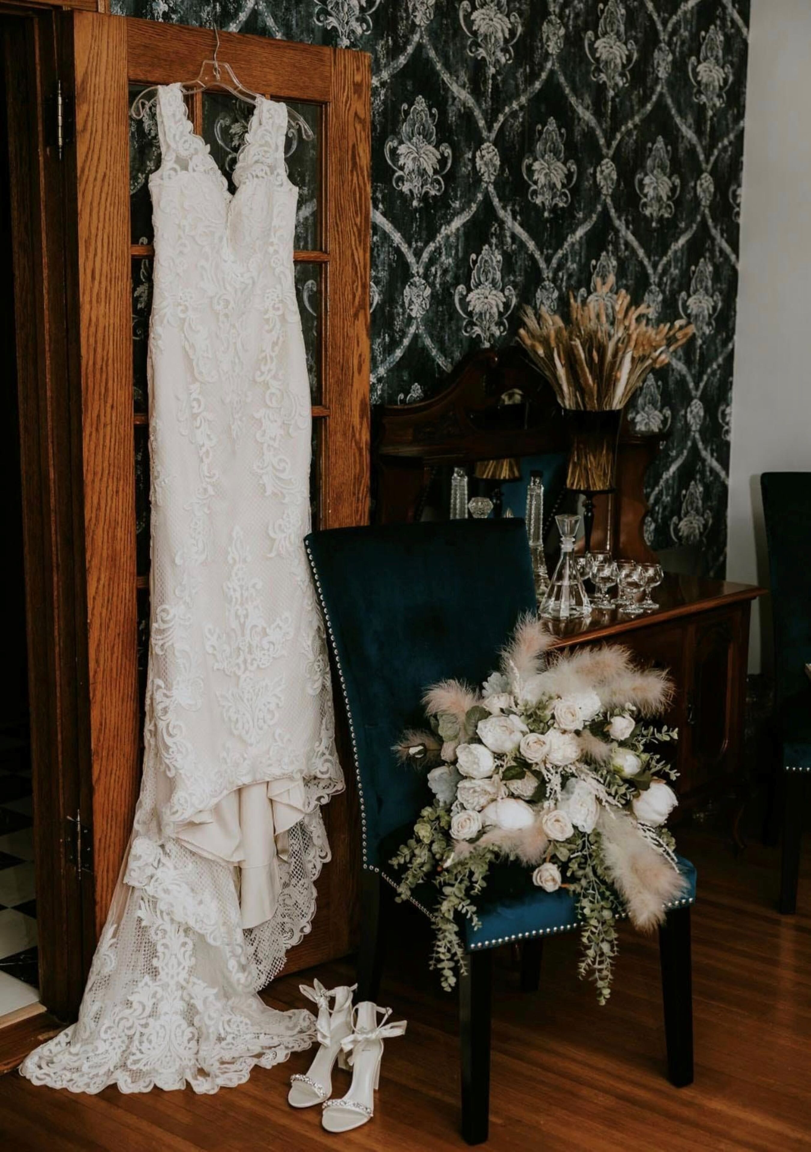 A wedding dress on a hanger, a chair with a floral arrangement, and a pair of shoes on the floor.