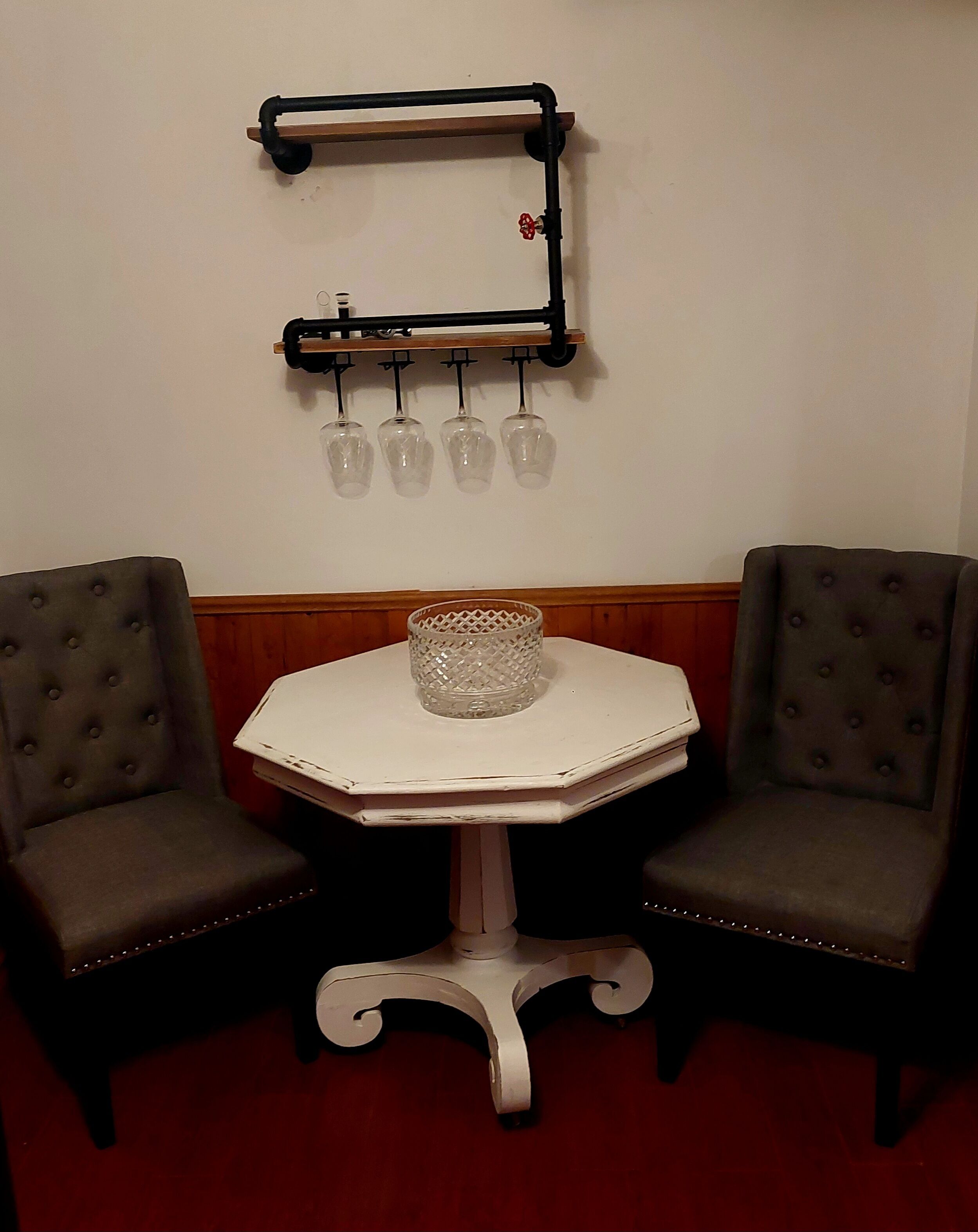 Una pequeña mesa octogonal con acabado blanco y un florero de vidrio en el centro. Dos sillones tapizados con detalles de botones están colocados a cada lado. Sobre la mesa, un estante de metal negro sostiene cuatro vasos de vidrio.