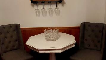 Una pequeña mesa octogonal con acabado blanco y un florero de vidrio en el centro. Dos sillones tapizados con detalles de botones están colocados a cada lado. Sobre la mesa, un estante de metal negro sostiene cuatro vasos de vidrio.