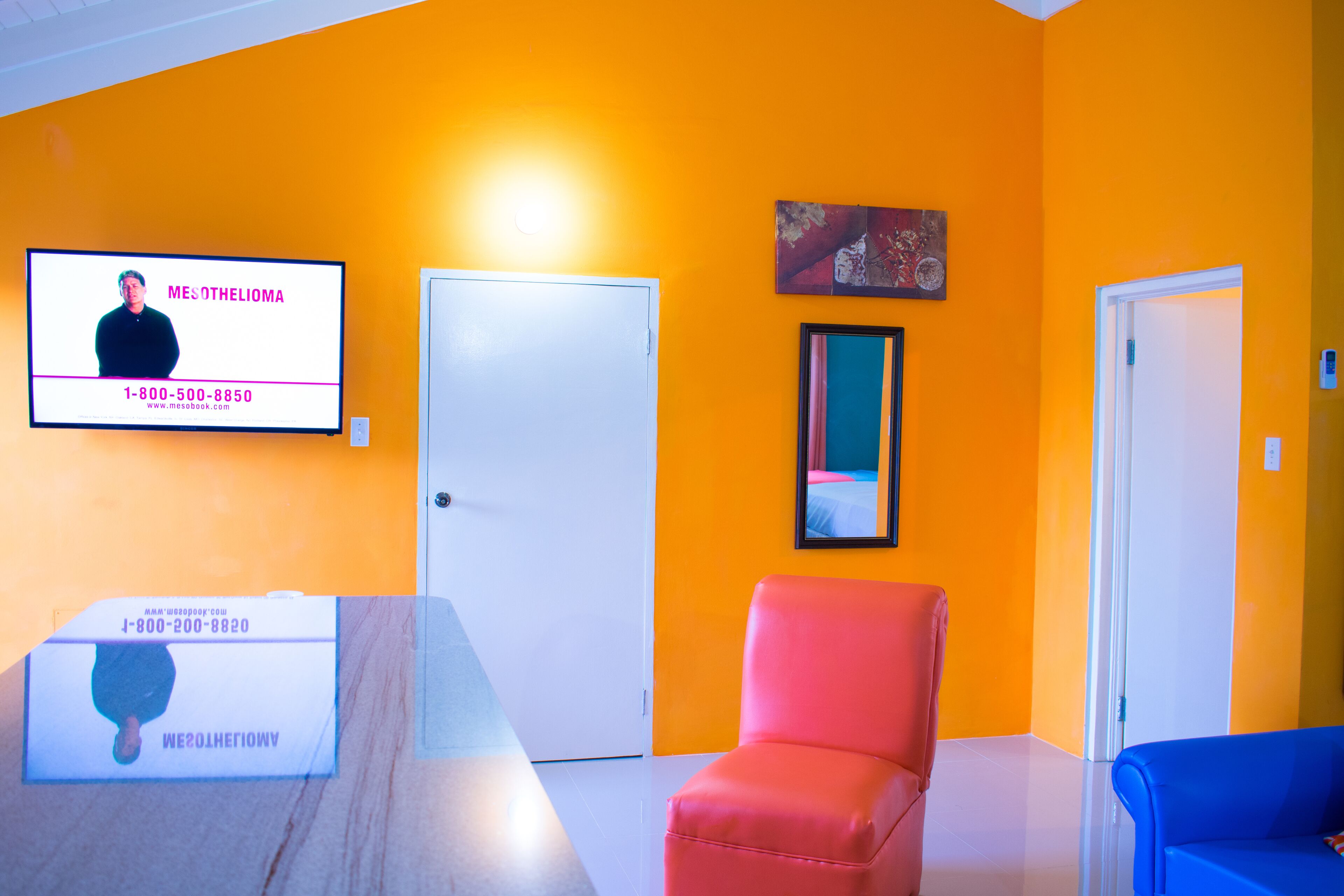 A room with a bright orange wall, a white door, a red chair, a blue sofa, and a television displaying a person with contact information.