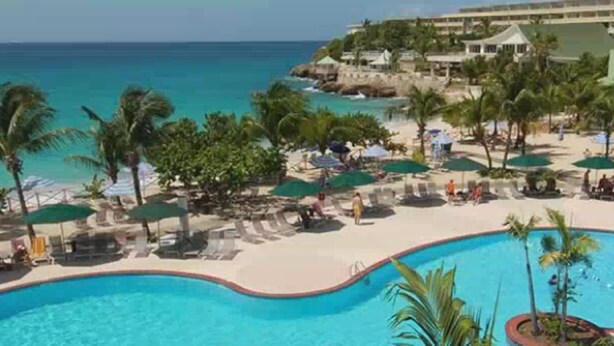 Zwembadgedeelte van een resort met uitzicht op de oceaan, palmbomen en een gebouw op de achtergrond.