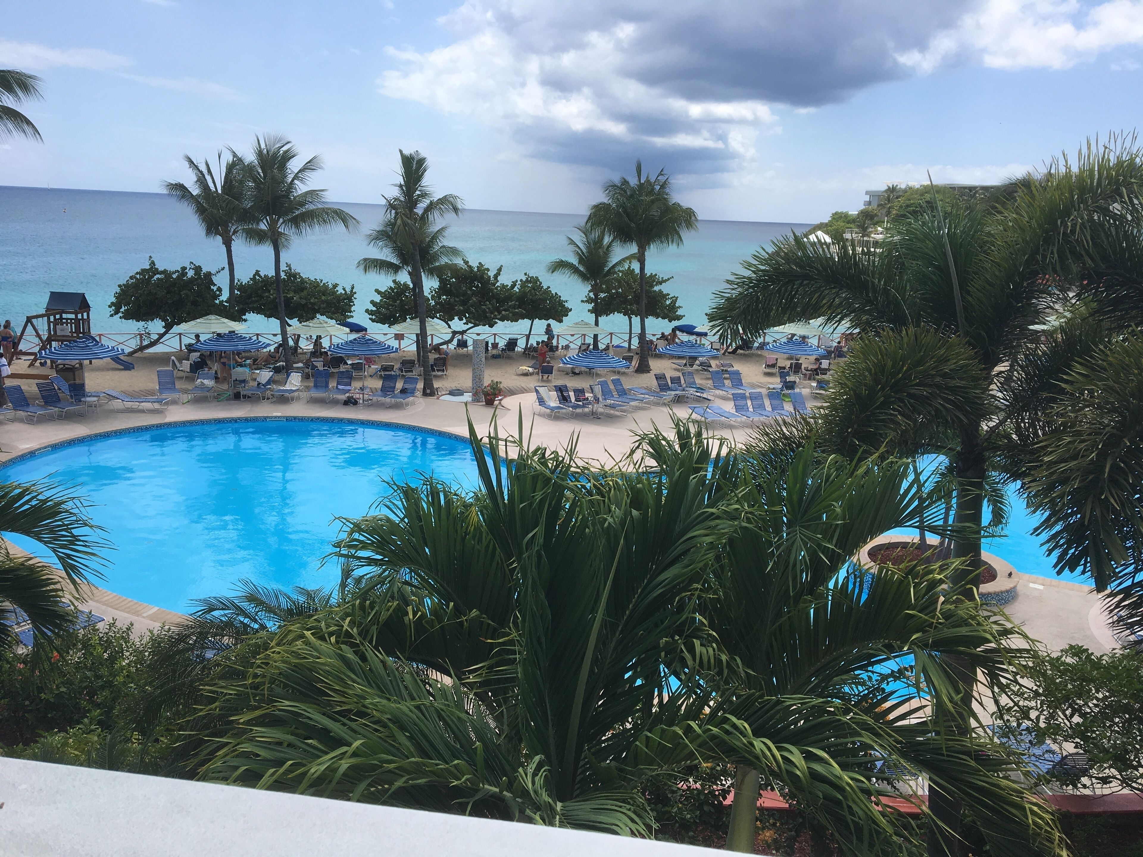 A poolside area with lounge chairs and umbrellas, a clear view of the ocean, and palm trees.