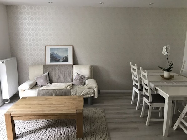 A living room with a sofa, a coffee table, a dining table with chairs, a framed picture on the wall, and a potted plant. - Villa Waldglück near Rangsdorfer See (Rangsdorf)