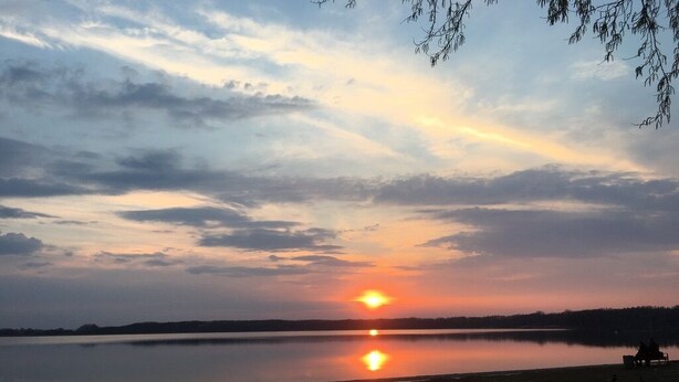 A sunset over a calm lake with a sandy shore and a few people sitting on the beach. - Villa Waldglück near Rangsdorfer See (Rangsdorf)