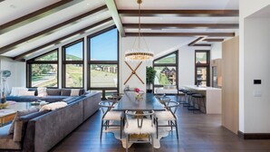 A spacious living area with a long dining table, a sofa, and large windows offering a view of the outdoors. - The Ultimate Summertime Luxury Penthouse Retreat in Snowmass Base Village! (Snowmass Village)