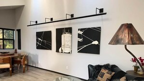 Una sala de estar moderna con comedor, que presenta una mesa de centro de vidrio, un sofá negro con una manta de piel y tres cuadros abstractos en la pared.