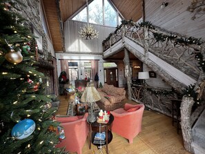 A spacious living area with a large Christmas tree, a cozy fireplace, and a rustic staircase.