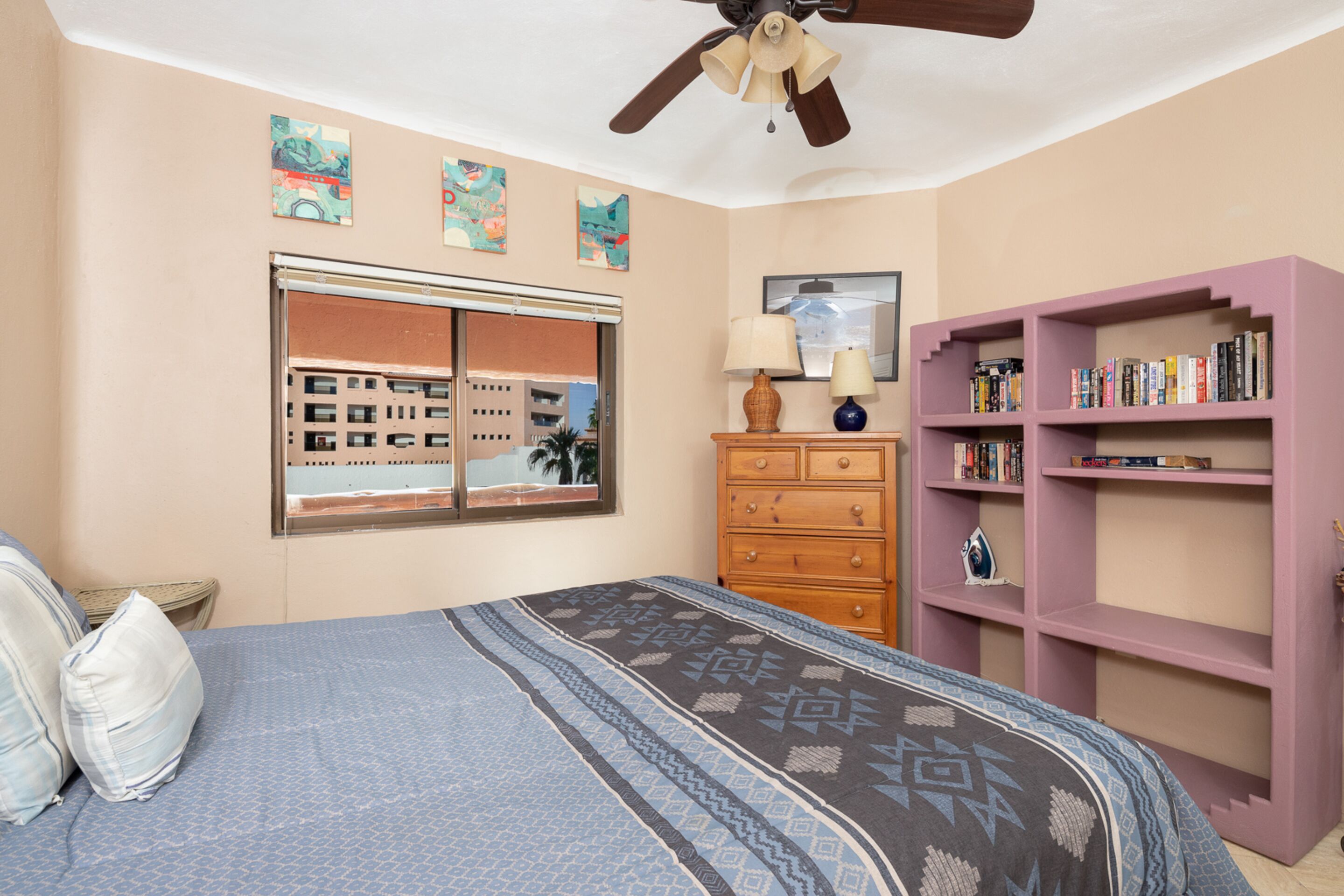 Ein Schlafzimmer mit einem Bett, einem Schminktisch, einem Bücherregal, einem Deckenventilator und Kunstwerken an den Wänden.