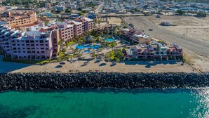 Vista aérea de un complejo turístico costero con varias edificaciones, una playa de arena y un litoral rocoso.
