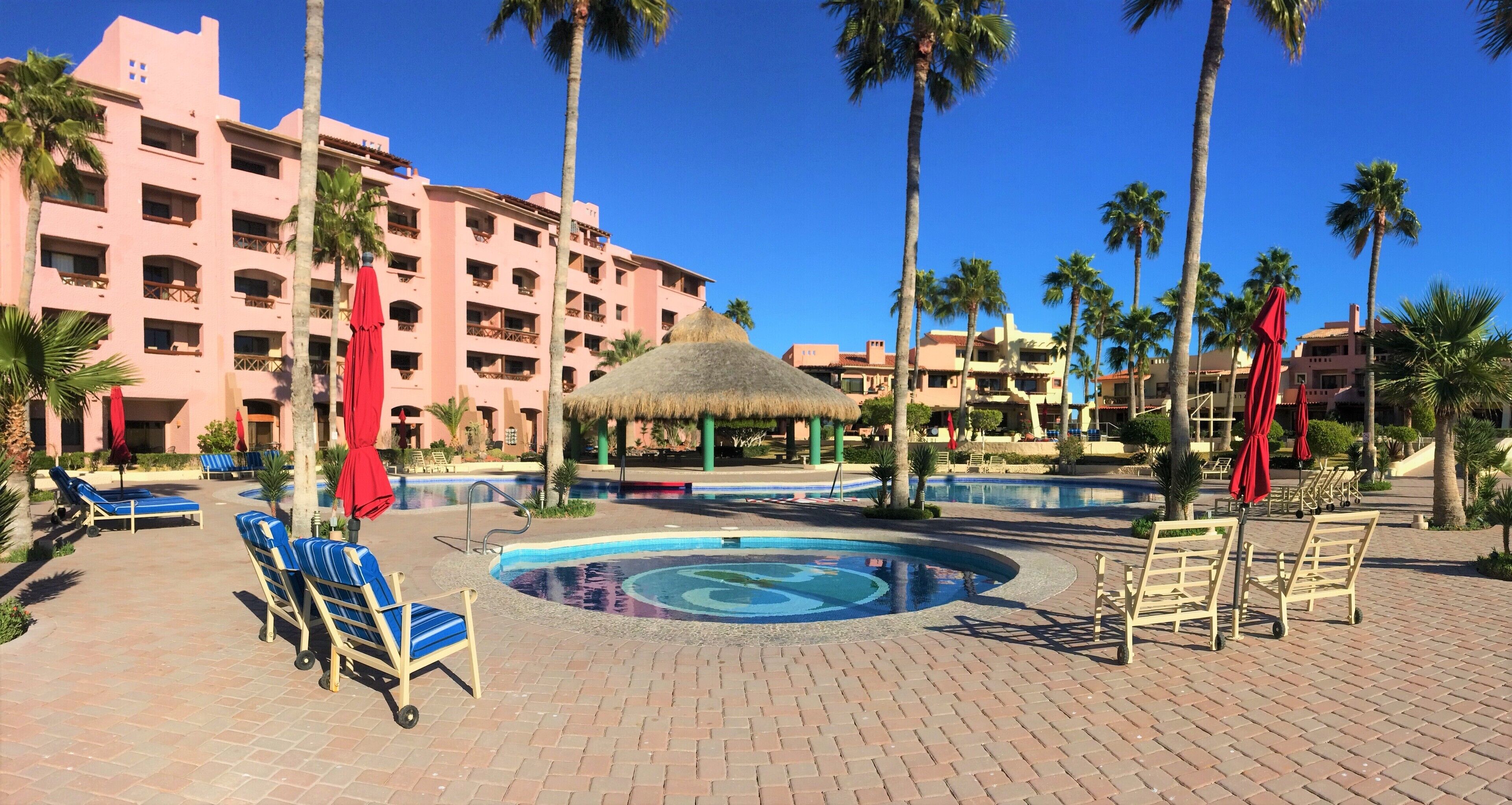 Zona de piscina de hotel con sillones de descanso, palmeras y un edificio grande al fondo.