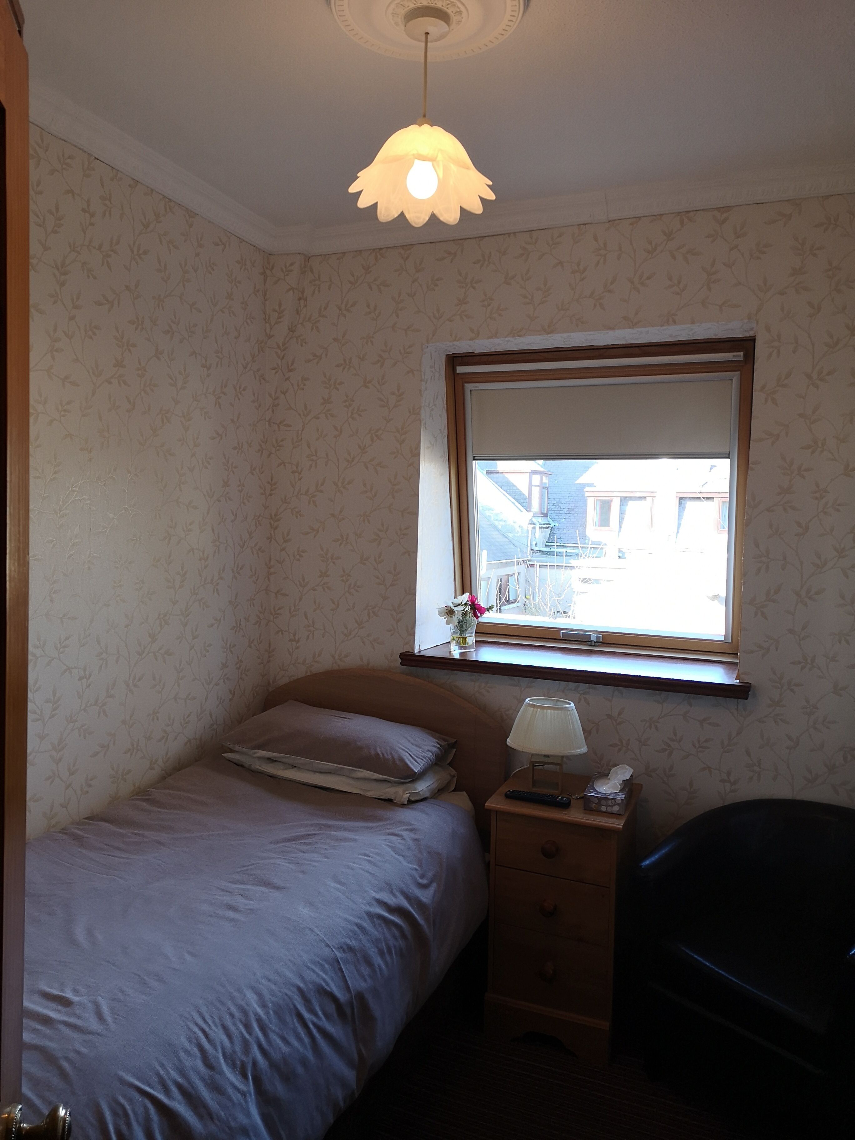 Un dormitorio pequeño y ordenado con una cama individual, una mesita de noche con una lámpara, una ventana con vistas a edificios y una lámpara en el techo.