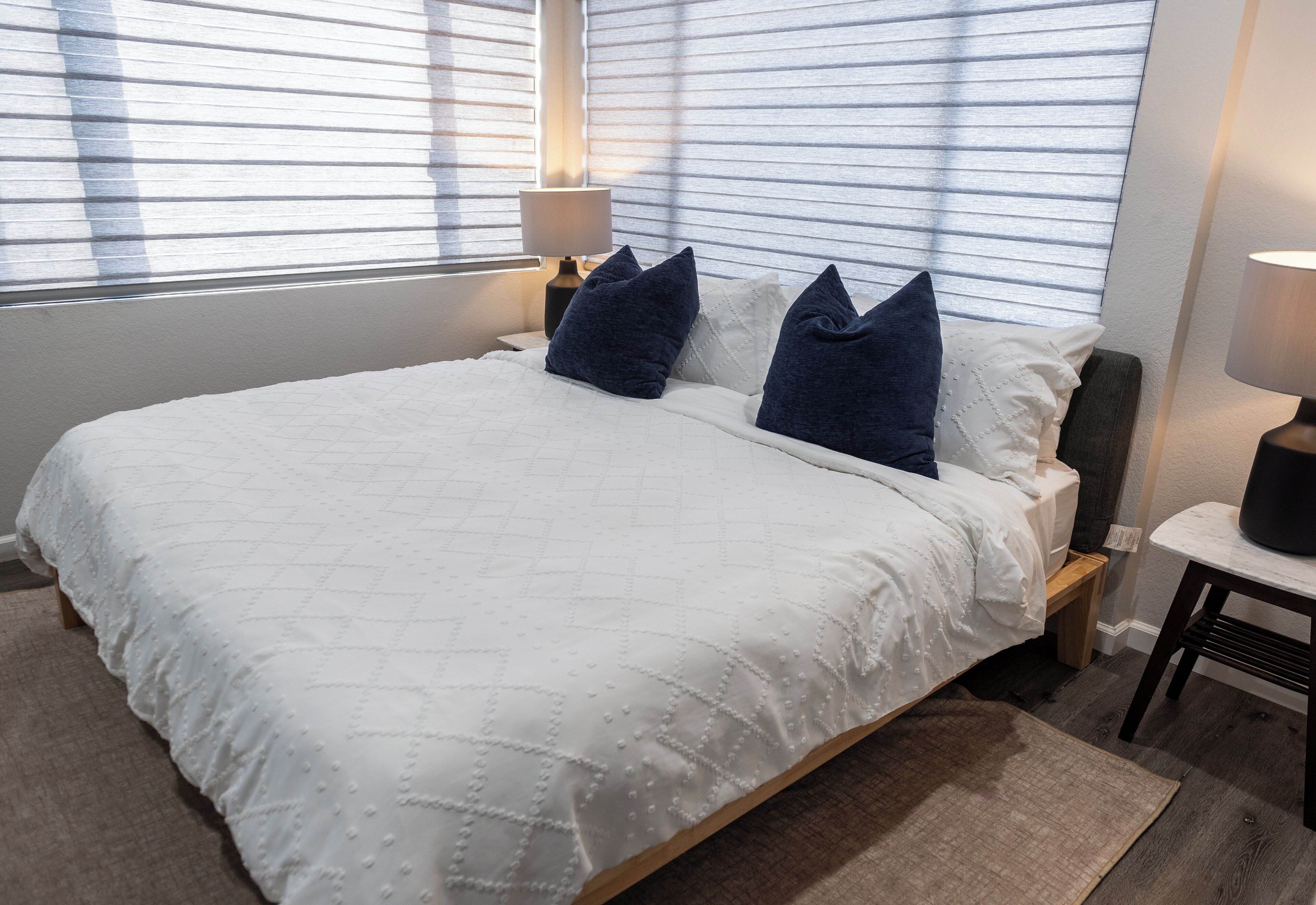 A neatly made bed with a white quilt, two dark pillows, and a bedside lamp.