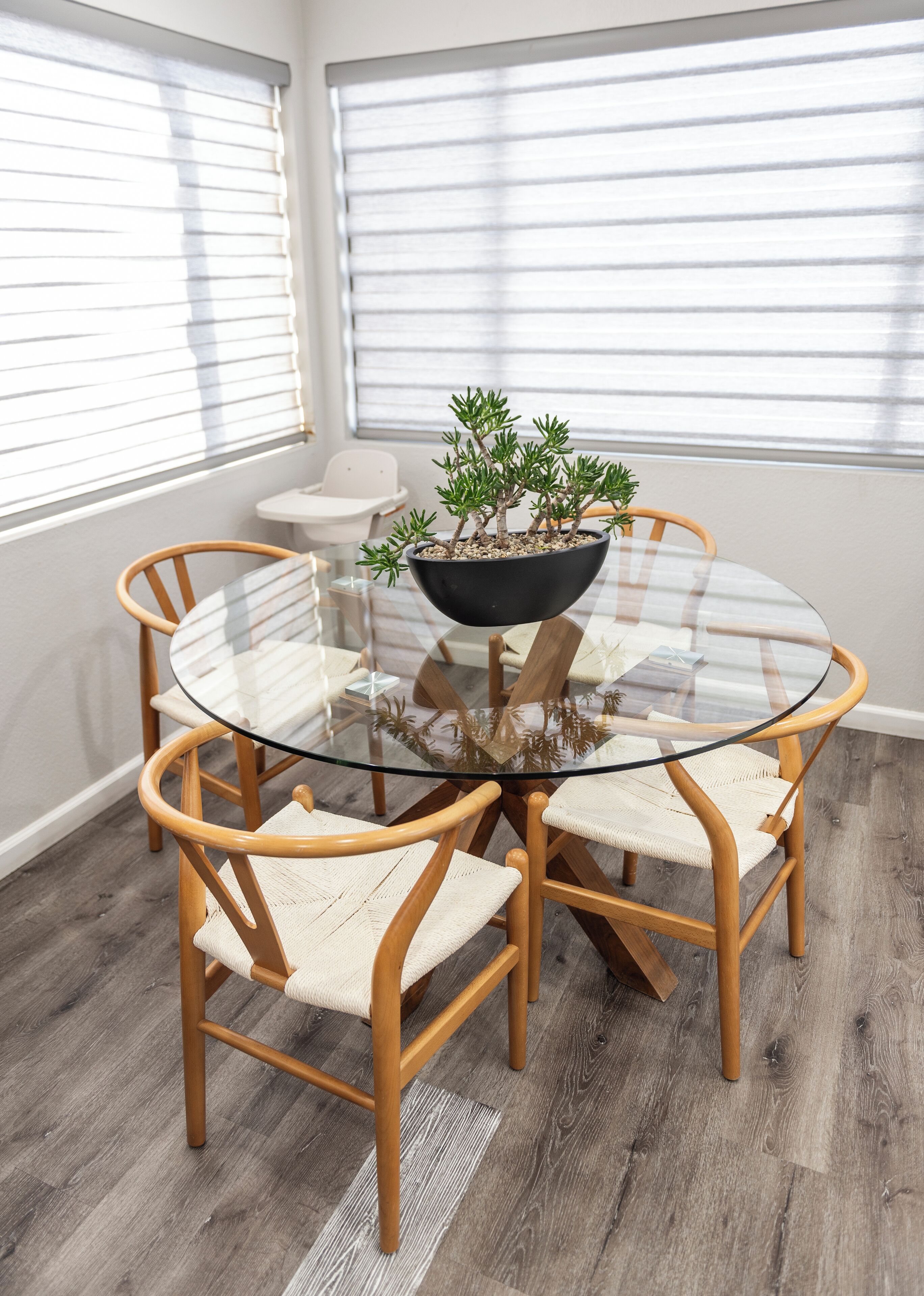 A round glass table with a black bowl containing a small plant, surrounded by wooden chairs with woven seats, in a room with wooden flooring and white walls.
