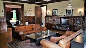 A spacious living room with a brown leather sofa, a glass coffee table, and a television set. There is a bookshelf filled with books and decorative items, and a dining area with a table and chairs is visible in the background. - Architect's Brownstone Mansion 6B6B, Porch, Garden (Minneapolis)