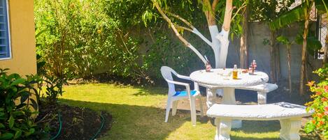 A garden with a round table and chairs, surrounded by greenery and a tree.