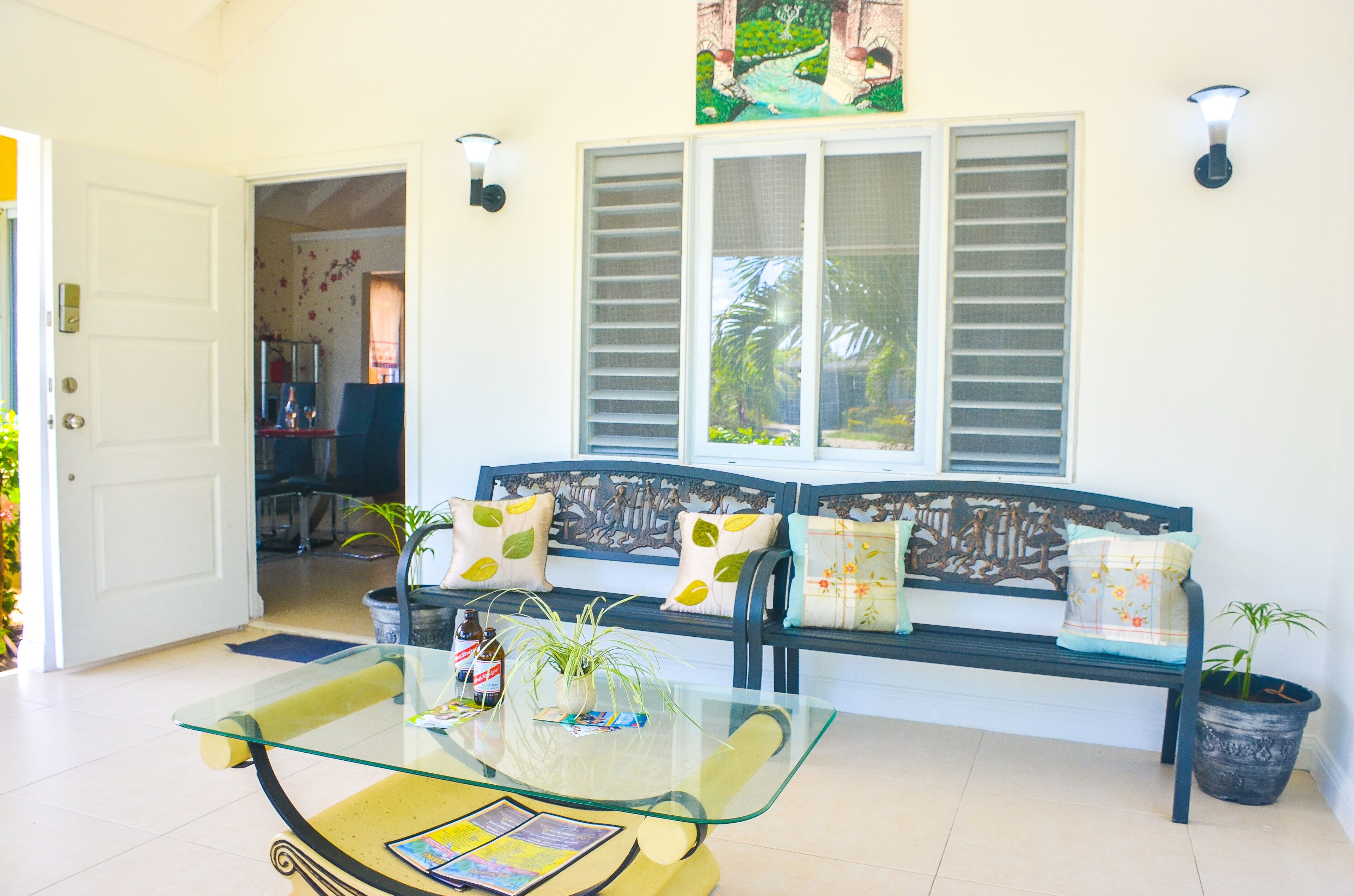 A living room with a glass coffee table, a sofa with patterned cushions, and a large window with shutters.